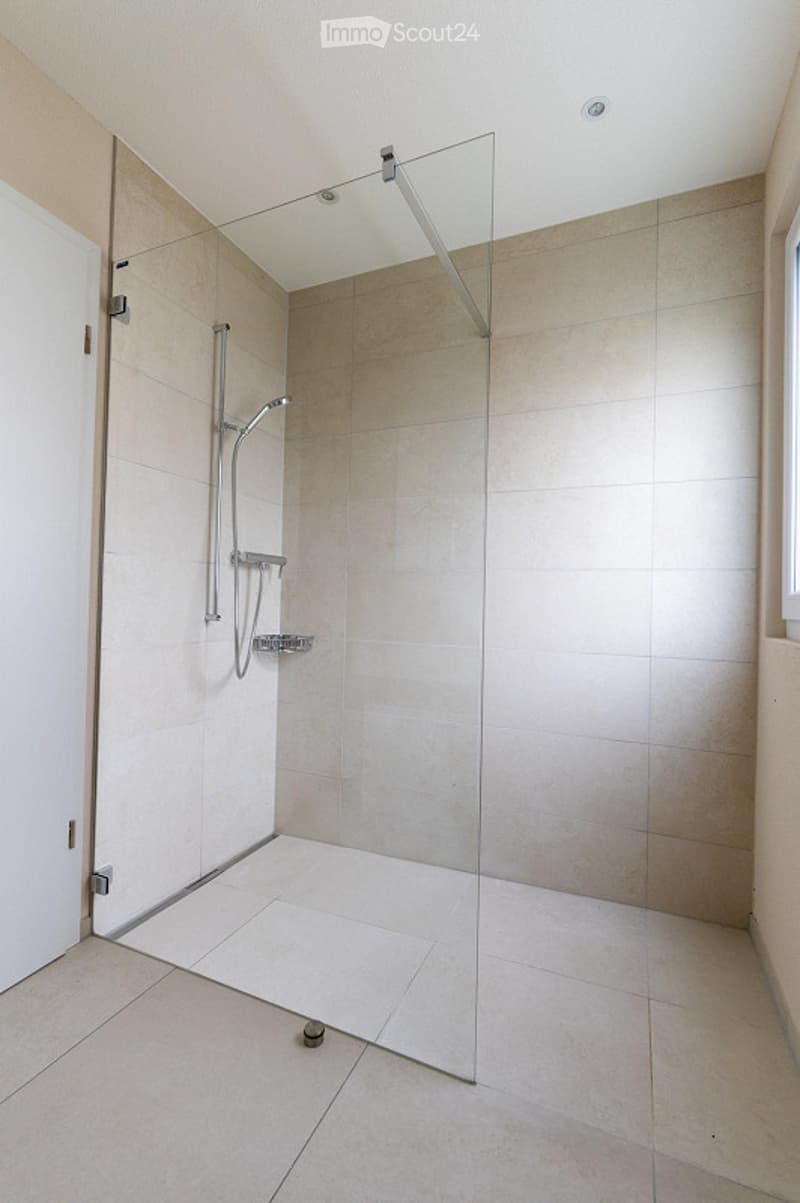 Modern bathroom with a large, clean shower stall featuring a glass door and a sleek shower handle. The walls and floor are tiled in neutral tones, creating a bright and contemporary look.