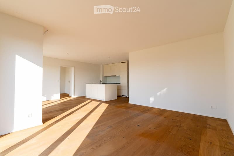 Bright, unfurnished room with wooden flooring and white walls. A small kitchen area with white cabinets and a countertop is visible in the background.