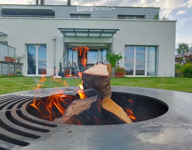 A fire pit with burning wood in the center of a circular metal grate, surrounded by a grassy area. Behind the fire pit, there is a modern white house with large glass windows and a patio with a table and chairs.