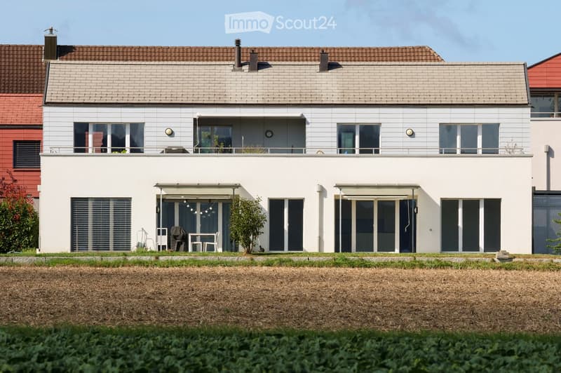 Modern two-story house with white exterior, grey roof, large windows, and a covered patio area with outdoor furniture.