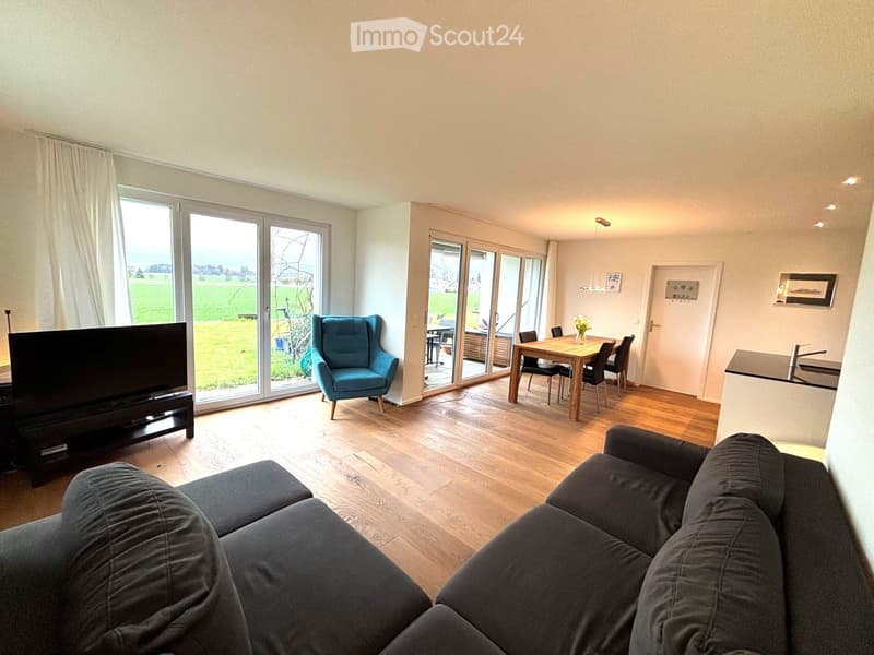 Living room with large windows, wooden floor, modern furniture, dining table, and a view of a green field
