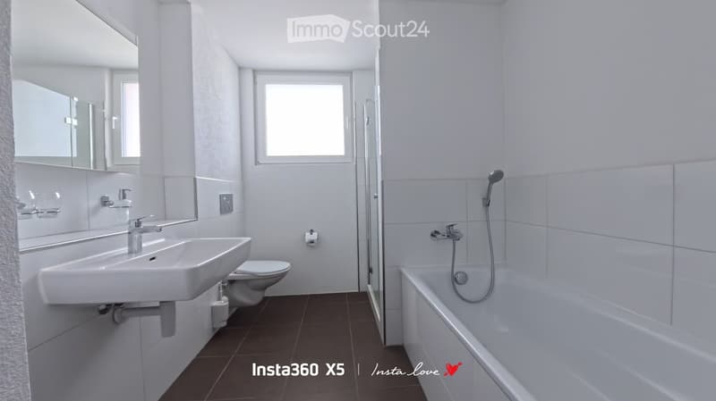 Modern bathroom with white tiles, a sink, toilet, bathtub, and a large mirror. The space is well-lit with a window providing natural light.