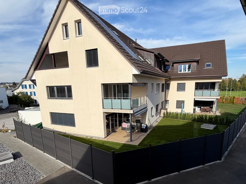 Modern two-story house with a balcony, solar panels on the roof, and a fenced garden area with patio furniture.
