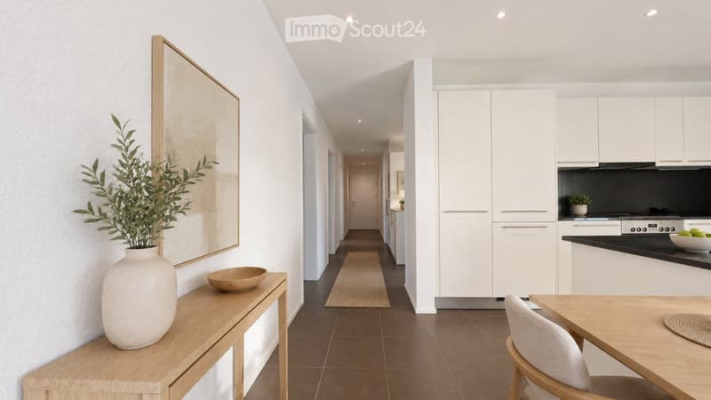 Modern hallway leading to a kitchen area with white cabinets, a black countertop, and a stove. A wooden table with a chair, a vase with greenery, and a bowl are placed in the hallway.