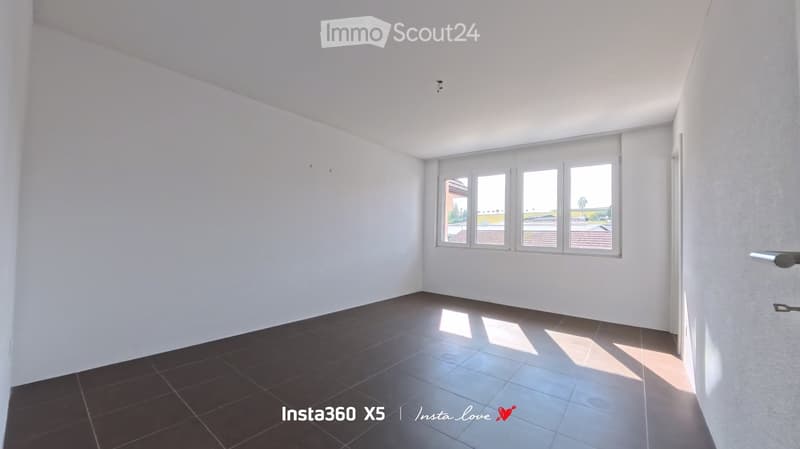 Empty room with white walls, dark tiled floor, and large windows letting in natural light.