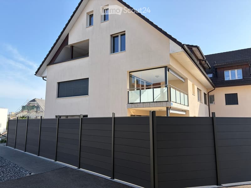 Modern two-story house with light beige facade, multiple windows, and a balcony with glass railing.