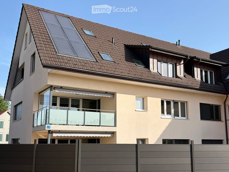 Two-story house with beige exterior, brown tiled roof, multiple windows, and a glass-enclosed balcony on the first floor.