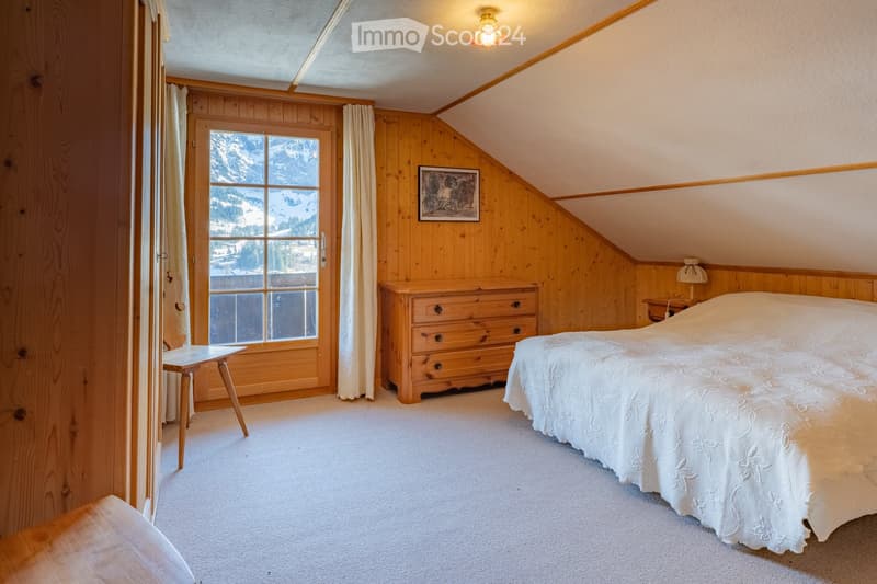 Cozy attic bedroom with wooden walls, slanted ceiling, large window offering a mountain view, bed, nightstand, chest of drawers, small table, and picture frame on the wall.