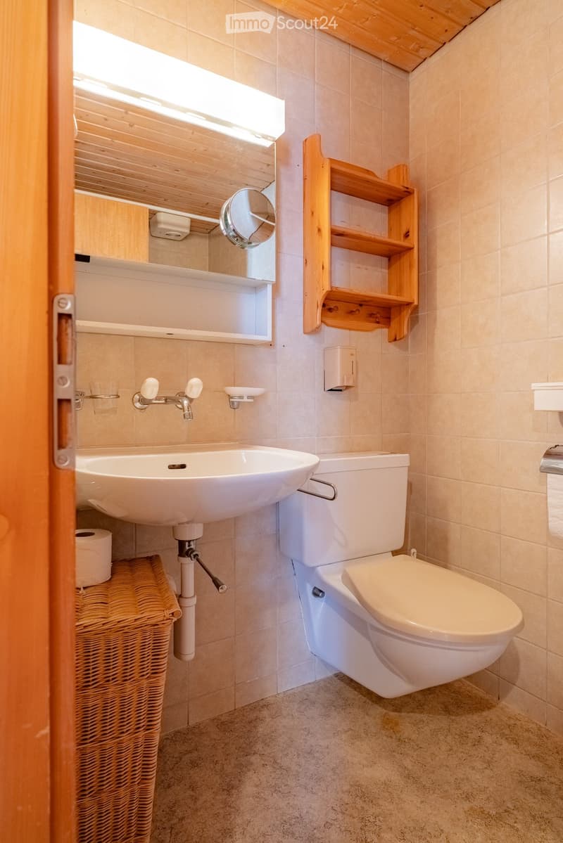 Modern bathroom with a white sink, toilet, mirror, wooden shelf, and wicker basket. Light-colored tiles and wooden accents.