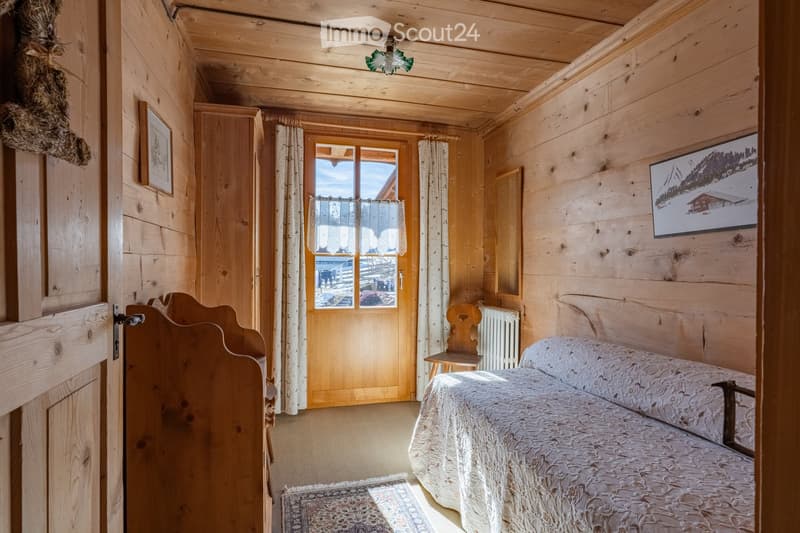 Cozy wooden bedroom with a single bed, a small wooden chair, a radiator, a picture frame, a TV screen, and a door leading to another room.
