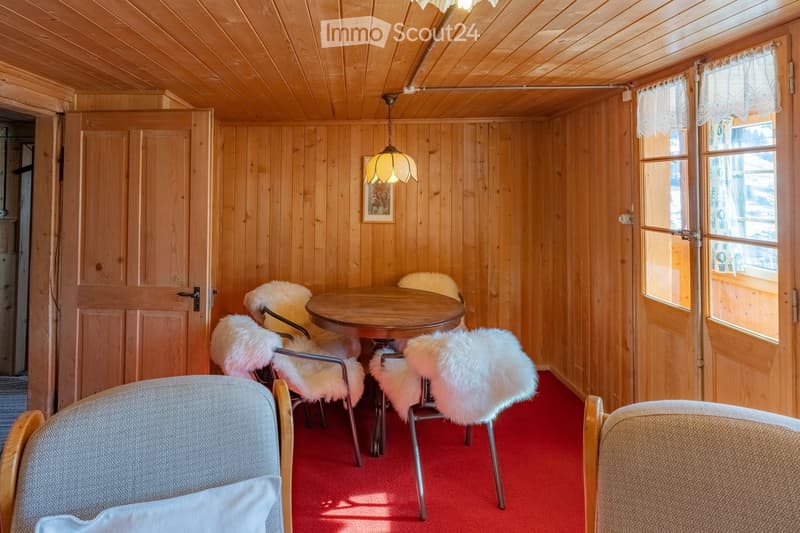 Cozy wooden room with red carpet, round wooden table, four chairs with fur cushions, picture frame on wall, and hanging lamp.