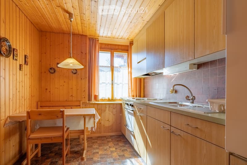 Cozy kitchen with wooden walls and ceiling, featuring a sink, oven, cabinets, a small dining area with a table and chair, and a hanging light fixture.