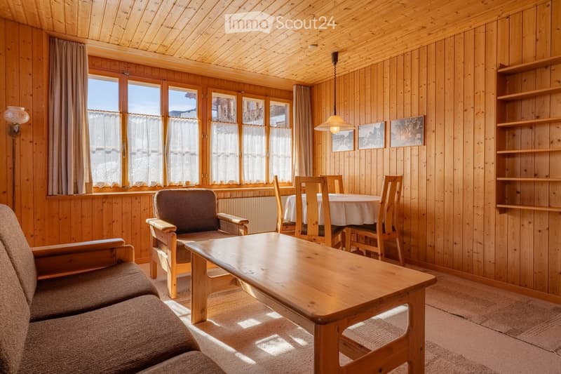 Cozy living room with wooden walls and ceiling, large windows with curtains, a sofa, coffee table, dining area with table and chairs, shelves, and artwork.