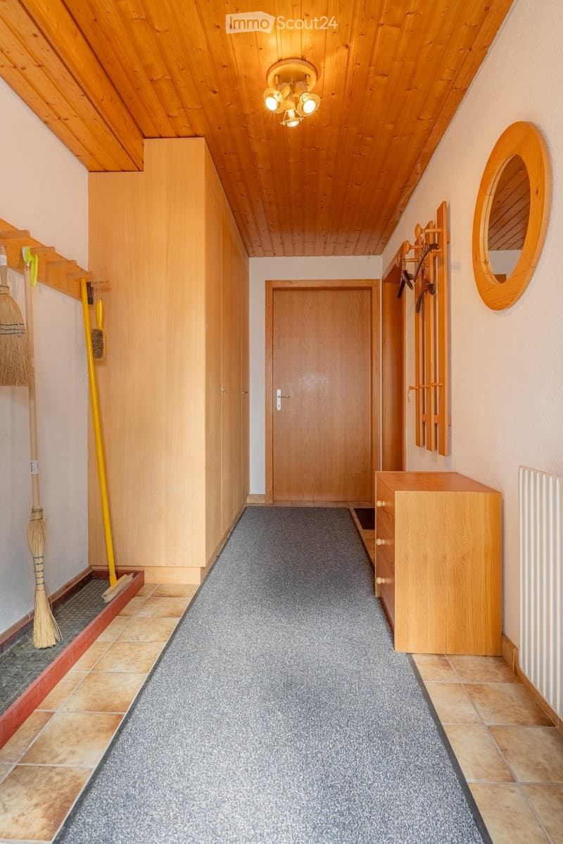 A narrow hallway with wooden walls, a grey carpeted floor, and a central wooden door. Features include a broom and dustpan on the left, a coat rack on the right, and a small wooden cabinet near the end.