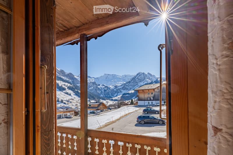 Balcony with wooden railings and a view of snow-capped mountains, a village, and parked cars below.