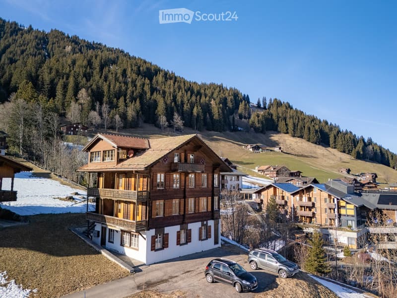 A large wooden house with multiple stories, balconies, and a red-tiled roof, situated in a snowy, forested area with other houses and parked cars nearby.
