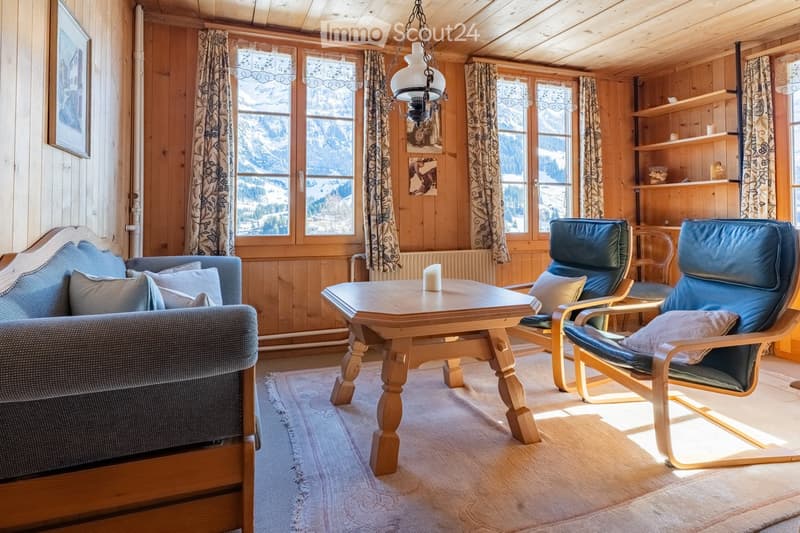 Cozy living room with wooden walls, large windows showing a snowy mountain view, a sofa, a wooden coffee table, two armchairs, and a shelf with decorative items.