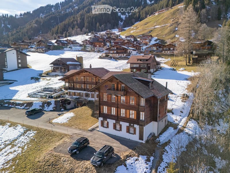 Aerial view of a large wooden house with multiple stories, surrounded by snow-covered ground and other houses in a village setting.