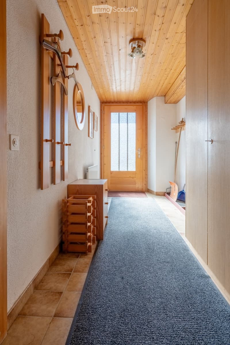 A narrow hallway with wooden ceiling beams, carpeted floor, and a wooden door at the end. Features include a coat rack, a small wooden cabinet, and a mirror on the wall.