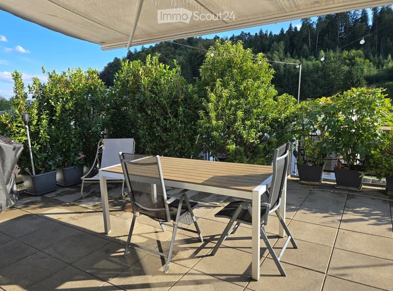 Balcony with a wooden table and four chairs, surrounded by lush greenery and a clear blue sky.