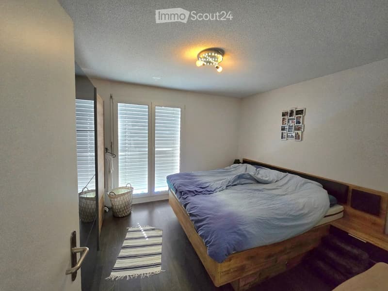Cozy bedroom with a wooden bed, blue bedding, a small rug, and two wicker baskets near the door. The room features large windows with blinds and a modern ceiling light.