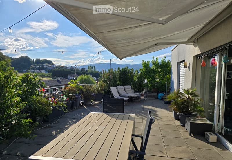 A spacious, covered balcony with wooden decking, lounge chairs, potted plants, and a scenic mountain view.