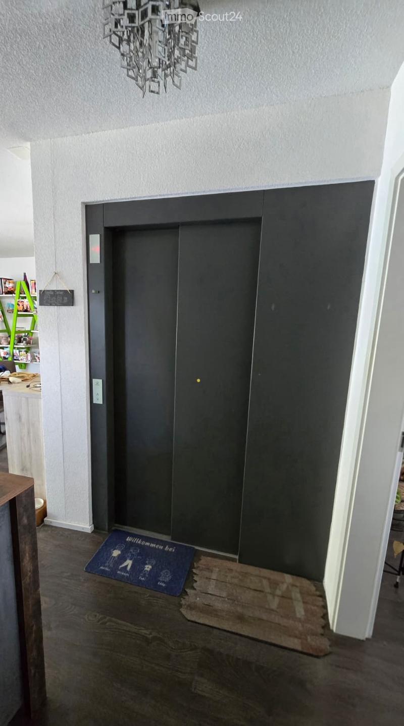 Modern hallway with dark wooden floor, white walls, and a black elevator door. A decorative rug with text is placed in front of the elevator.