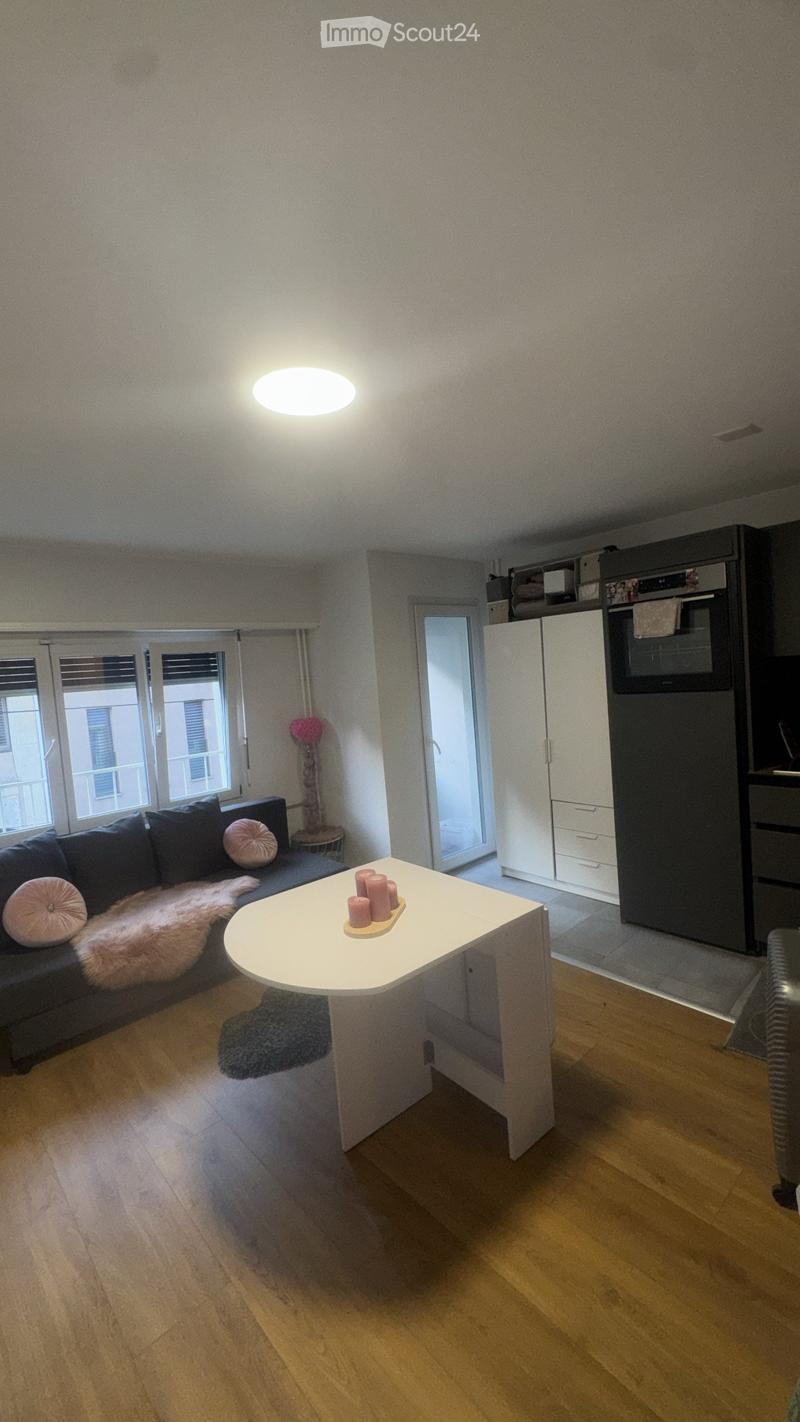 Modern living room with a dark blue sofa, white coffee table, and a small kitchen area with white cabinets and black appliances.