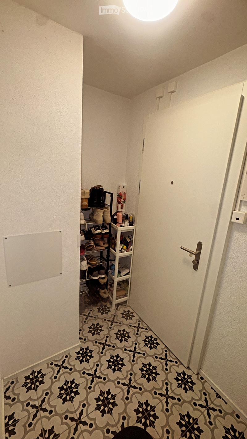 A narrow hallway with patterned tile flooring, white walls, and a small shoe rack holding various shoes and bags. A closed door is visible on the right side.
