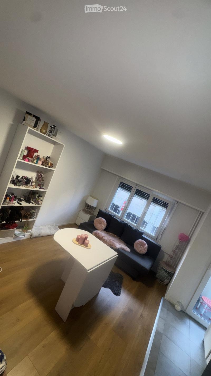 A modern living room with wooden flooring, a black sofa with pink cushions, a white coffee table with candles, a shelf with various items, and a lamp on a side table.