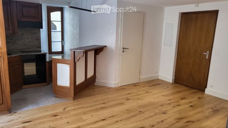 Empty room with wooden flooring, white walls, and a wooden bar counter. There are two doors and a small kitchen area visible in the background.