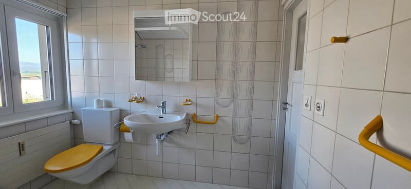 Bathroom. Modern bathroom with white tiles, a toilet with a yellow seat, a sink with a mirror cabinet, and yellow handrails for accessibility.
