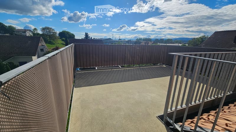 Balcony / Terrace. Roof terrace with concrete surface, metal railing, and wooden decking in the corner. The view includes neighboring houses, trees, and distant mountains under a partly cloudy sky.