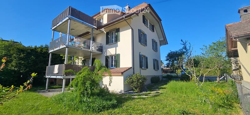 House. Two-story house with beige walls, green shutters, and a large balcony on the upper floor. The house is surrounded by a well-maintained garden with grass and shrubs.