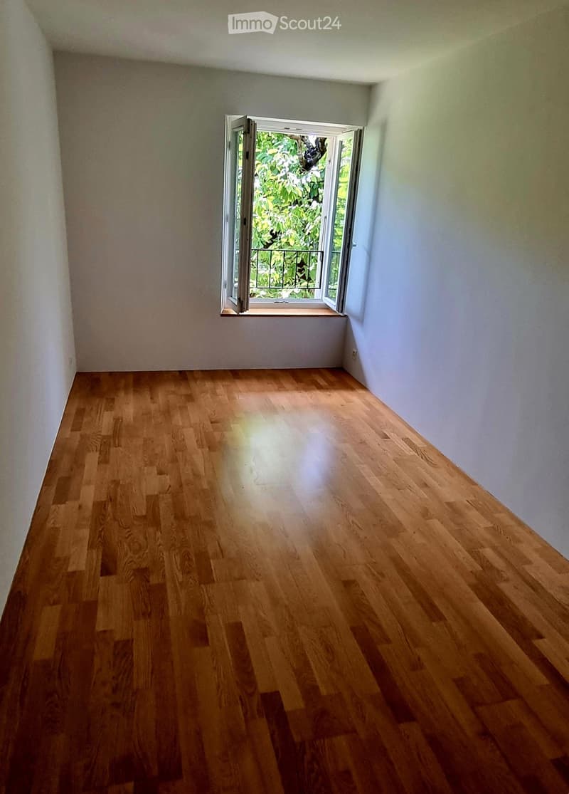 Empty room with wooden flooring and white walls, featuring a large window with a view of greenery outside.