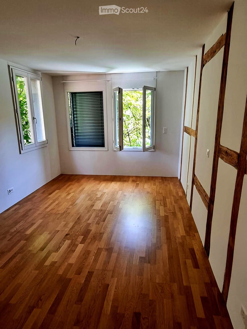 Empty room with wooden flooring, white walls, and large windows with blinds. The space is well-lit and has a simple, clean design.