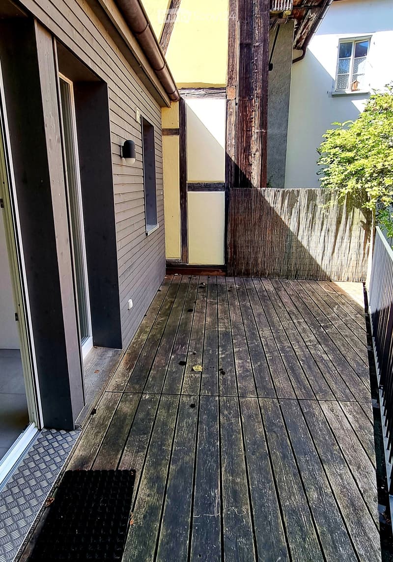 Wooden balcony with a wooden deck, white railing, and adjacent building with a window and greenery.