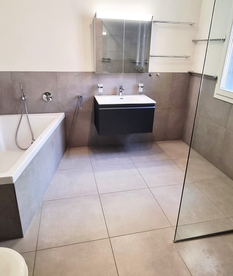 Modern bathroom with a bathtub, sink, and large mirror. The space features tiled walls and floor, with a glass shower enclosure visible.
