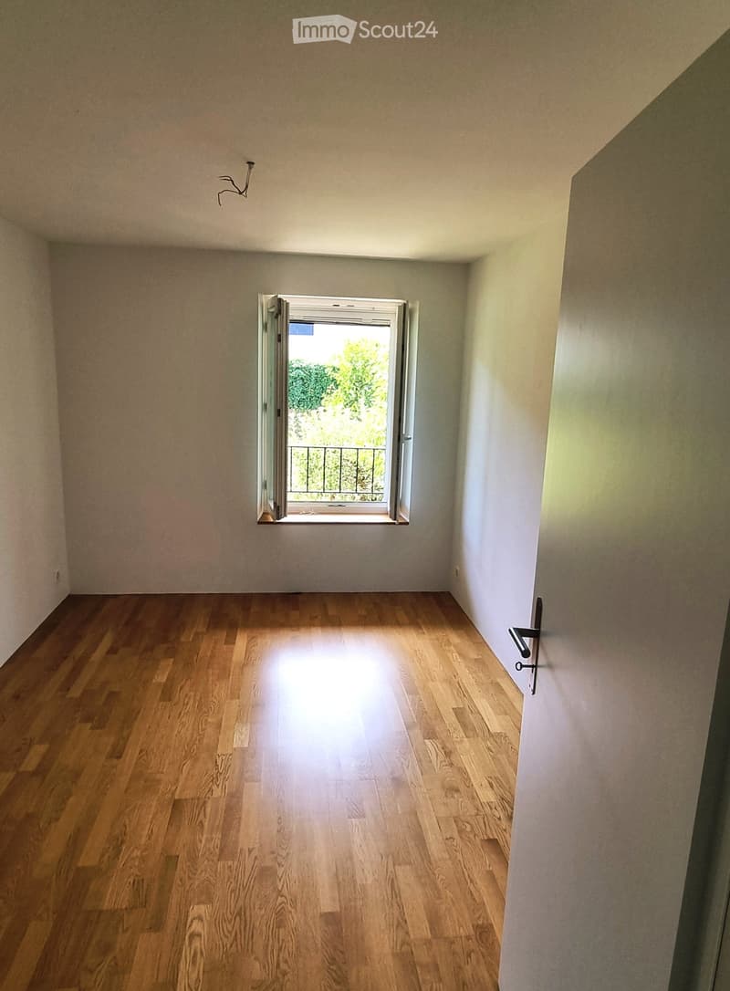 Empty room with wooden flooring and white walls, featuring a large window with a view of greenery and a balcony outside.