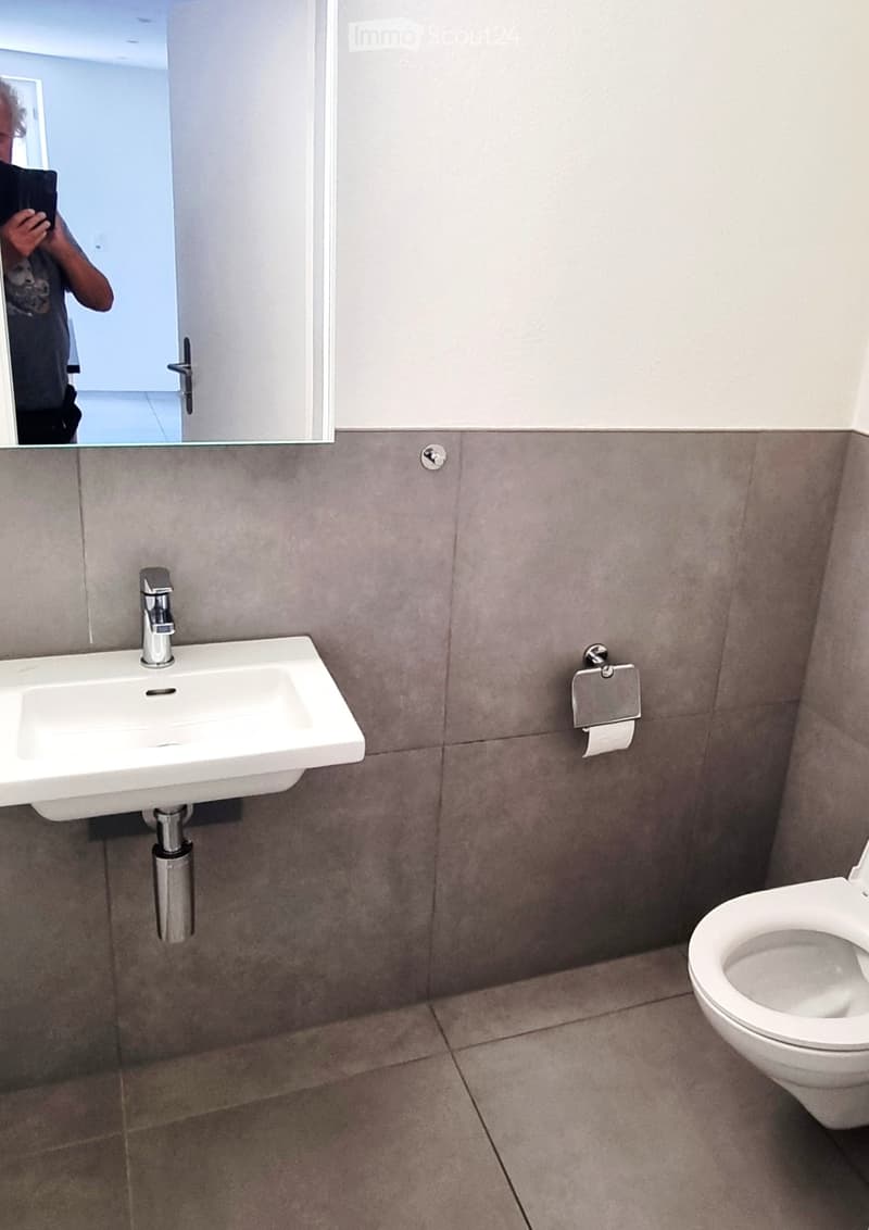 Modern bathroom with a white sink, chrome faucet, toilet, and a large mirror above the sink. Grey tiled walls and floor.