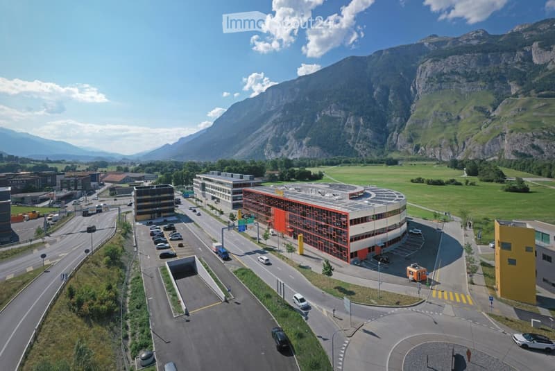 Aerial view of a modern building complex with a parking lot, surrounded by green fields and mountains in the background.