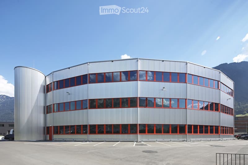 Modern, curved building with a metallic facade and large red-framed windows, surrounded by a parking lot and mountains in the background.