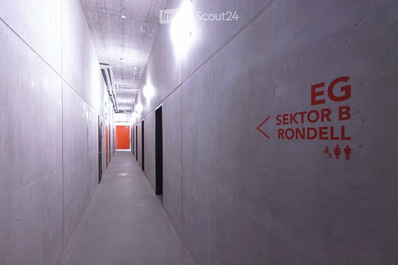 Modern, clean corridor with white concrete walls and ceiling, illuminated by recessed lighting. Signage indicates directions to 'EG SEKTOR B RONDELL' and includes accessibility symbols.