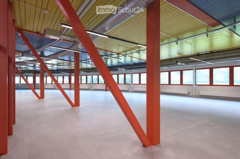 Large, open, industrial-style space with red structural beams, exposed ductwork, and large windows. The floor is smooth and light-colored, and the ceiling has a yellow panel design with integrated lighting.