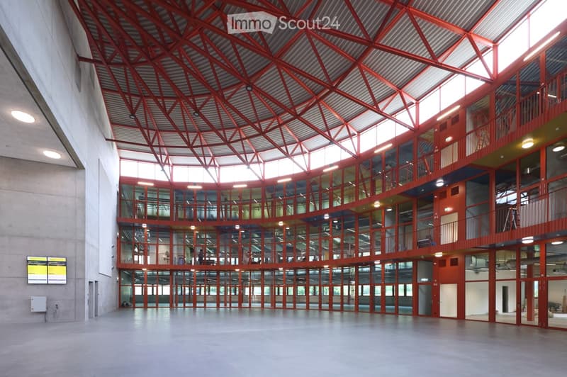 Modern interior space with a high, angular red metal ceiling and large glass walls, featuring multiple levels with open walkways and bright lighting.