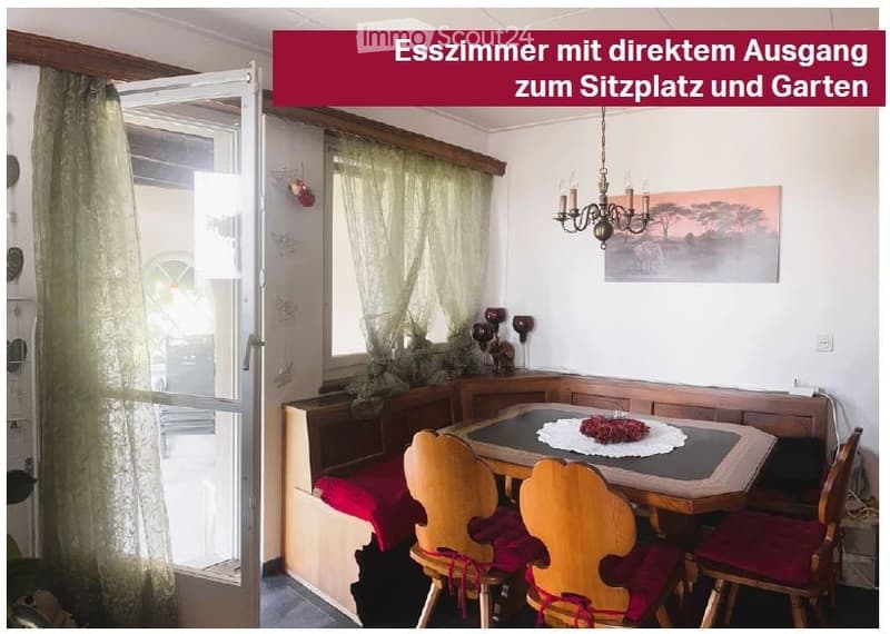 Dining room with a wooden dining table, four wooden chairs, a built-in bench with red cushions, a chandelier, and a picture frame on the wall. There is a large window with curtains and a door leading to an outdoor seating area.