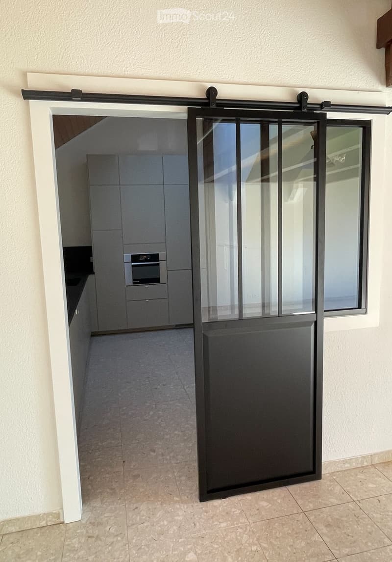 A modern sliding glass door with a black frame and glass panels, featuring a sleek design and a smooth, tiled floor in the hallway.