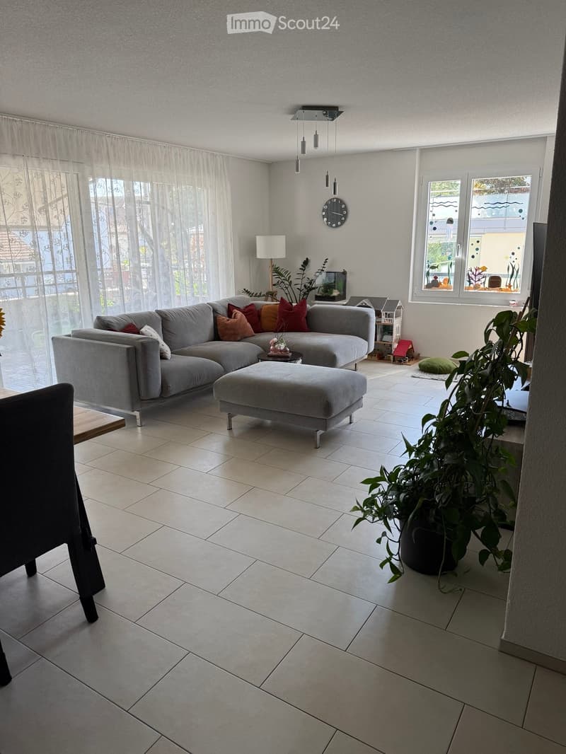 Modern living room with a grey sectional sofa, ottoman, small table, and decorative pillows. Large windows with sheer curtains, pendant lights, and a potted plant near the window.