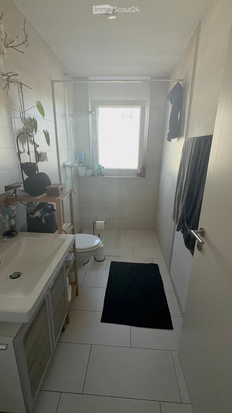 Modern bathroom with white tiles, a sink, toilet, shower area, and a small shelf holding various items. A black bathroom mat is placed in front of the toilet.