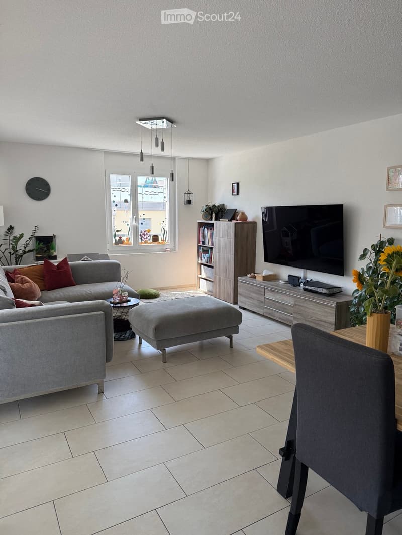 Modern living room with grey sofa, ottoman, coffee table, and a television mounted on the wall. The room features a bookshelf, a small table with a plant, and a large window with a view.